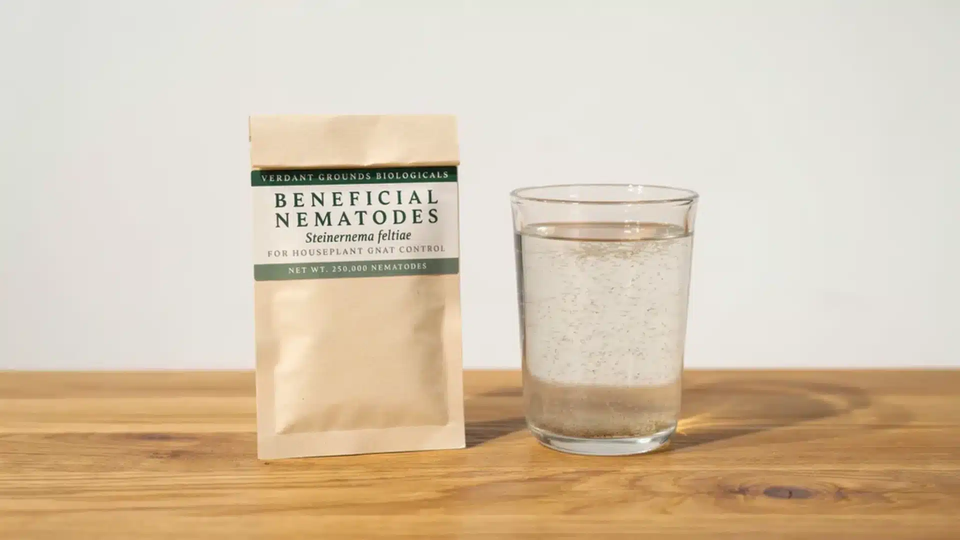 Package of beneficial nematodes next to a glass of water on a wooden surface for pest control use.